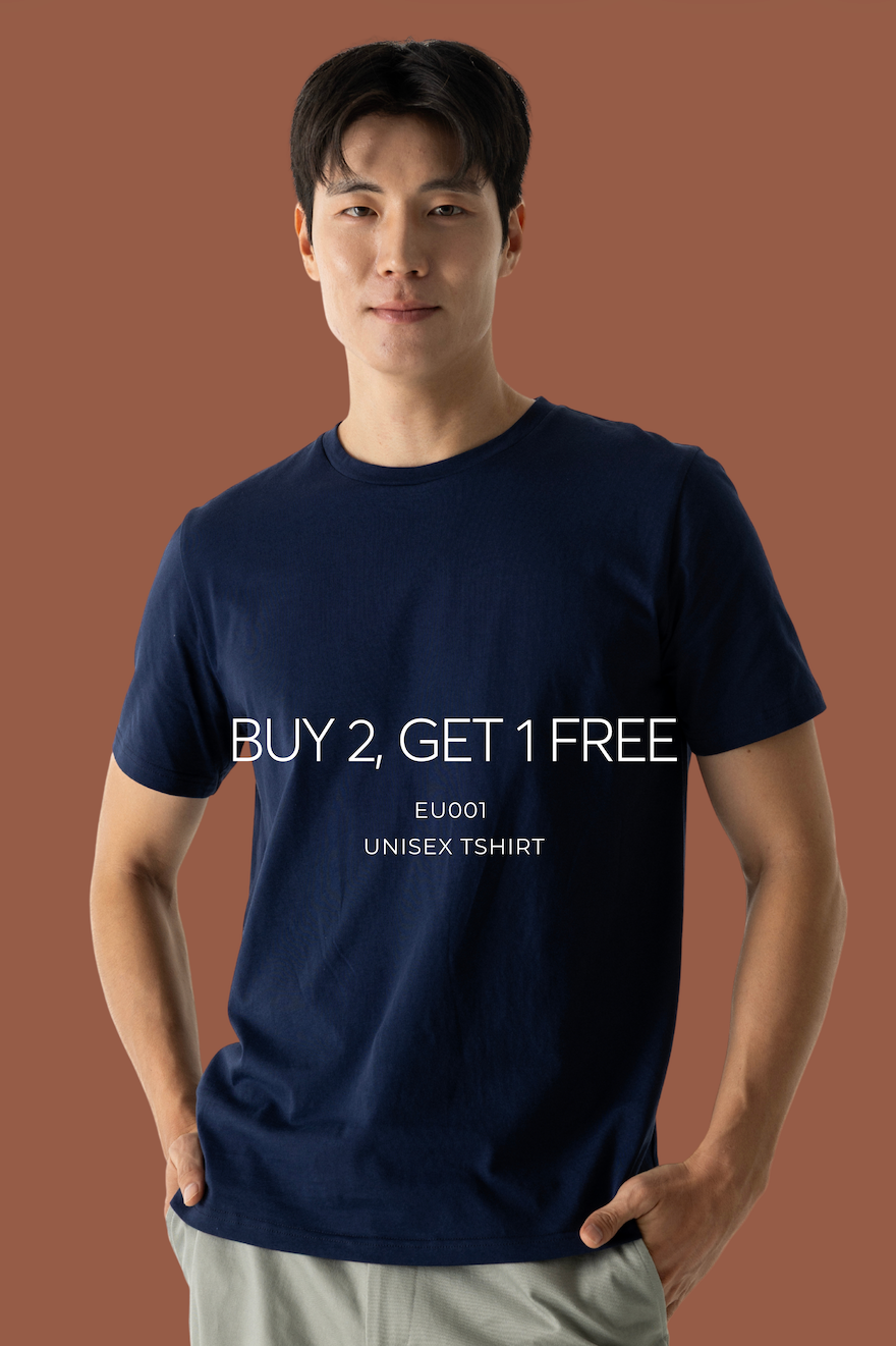 A man wearing a navy blue crewneck t-shirt with white text that reads 'BUY 2, GET 1 FREE' and 'EU001 UNISEX TSHIRT' on the front, paired with light gray pants, set against a solid terracotta background.
